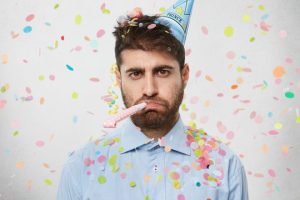 Man with a party hat and party whistle showered with confetti and looking sad.