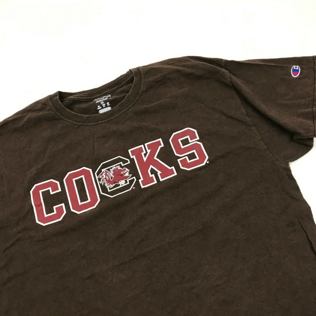 Brown t-shirt with the word "cocks" written in a bold football font in red with white outline. The middle character has no red fill, instead a picture of a red rooster is inserted inside the "c".