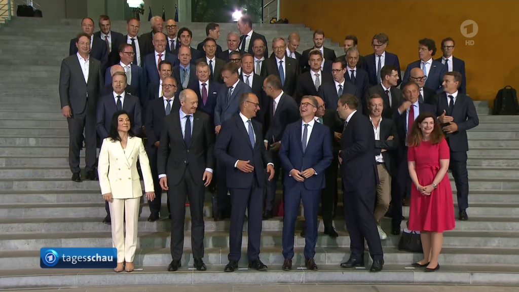 Screenshot of German nightly news, in which the chancellor is posing with approximately fifty CEOs. Only two of them are female, prominently positioned. The rest are all wearing suits. Two are bold enough to not wear ties. Presumably those are communists who slipped past security.
