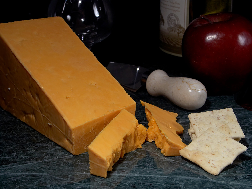 A cheese board with a large wedge of red leicester and some crackers.  In the background a red apple is being held by an invisible velociraptor.