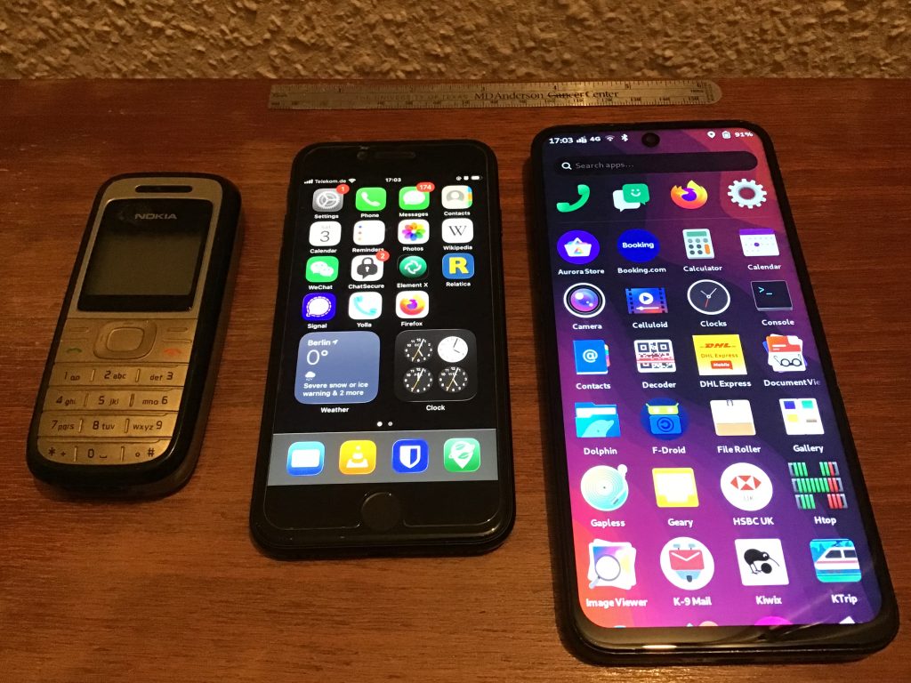 Three phones laid out side-by-side on a wooden bench.  There is a Nokia 1200 from 2008, an iPhone SE, and an FLX1s.  The last two are switched on and showing their home screens.  A small metal ruler is at the back for scale.