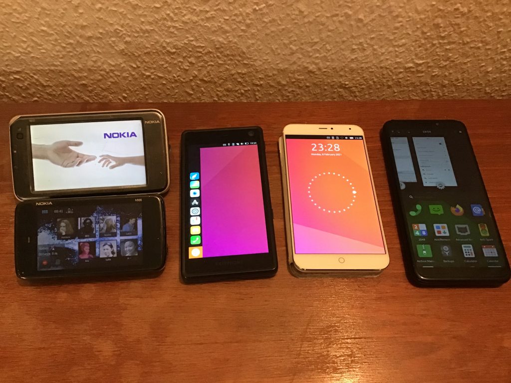 Several phones lined up on a wooden bench.  Left to right, a Nokia N810, a Nokia N900, a Fairphone 2, a Meizu MX4 Pro, and a Pinephone.  All the phones are switched on.  The N810 shows the Nokia reaching hands image.  The N900 shows a home screen with several contacts.  The Fairphone 2 shows the Ubuntu menu.  The Meizu MX4 shows the Ubuntu lock screen.  The Pinephone shows open apps at the top and a grid of icons at the bottom.