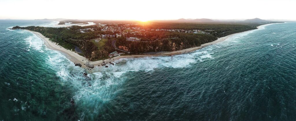 Aerial photograph of a spit of land at sunset, surrounded by beaches with heavy surf, and many buildings of a small town visible further away.